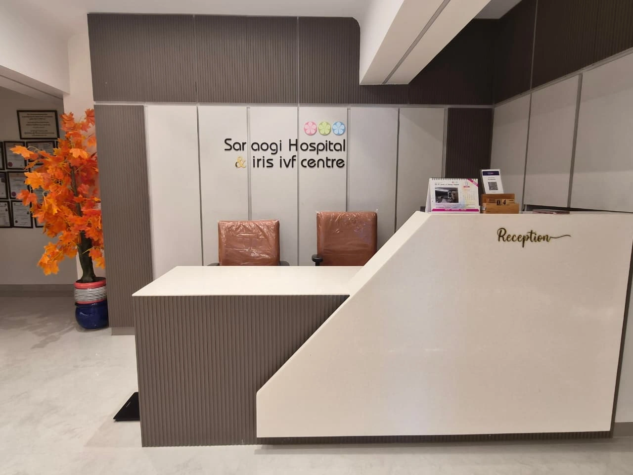 Hospital reception area with a white angled front desk, two brown chairs behind it, and a wall sign reading 'Sar aogi Hospital & iris IVF centre'; orange plant on the left.