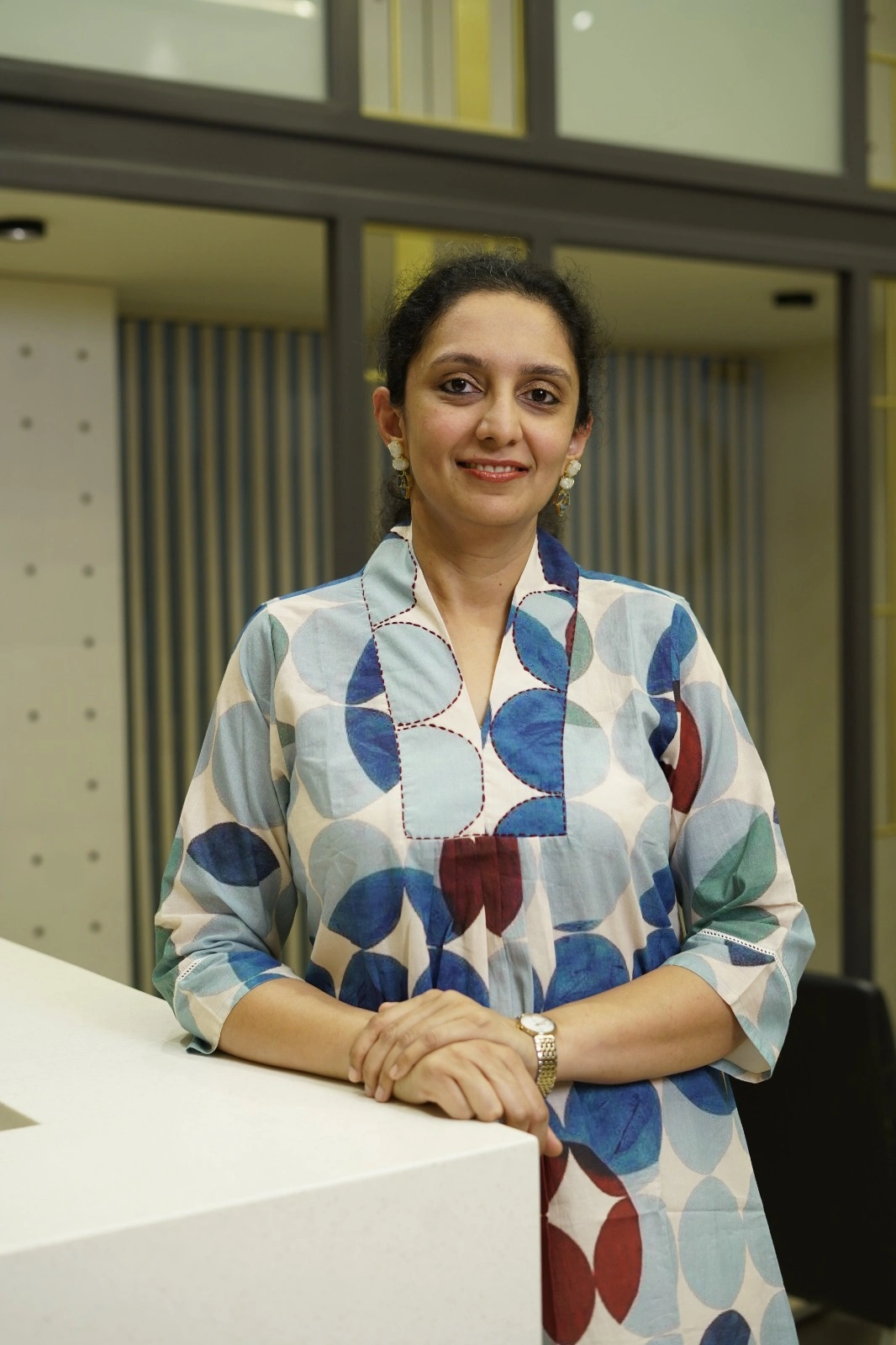 Smiling woman in a blue circular-patterned kurti standing behind a white counter in an office setting, hands folded on the counter.