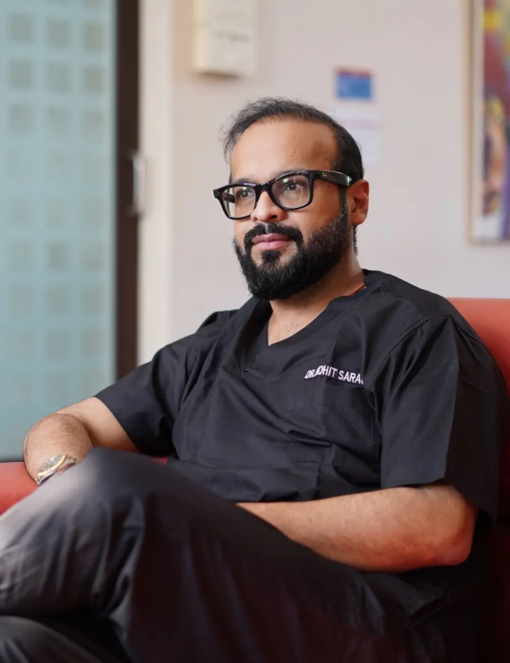 Male doctor with a beard and glasses sits in a clinic lounge wearing black scrubs.