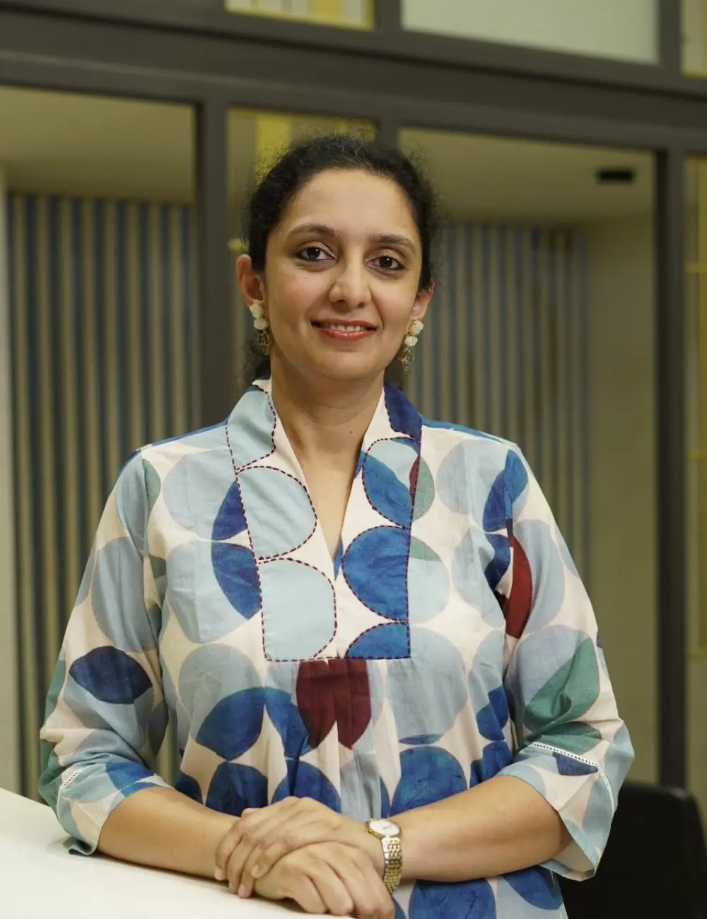 Smiling woman with dark hair wearing a blue-teal circular-patterned blouse, seated at a table indoors.