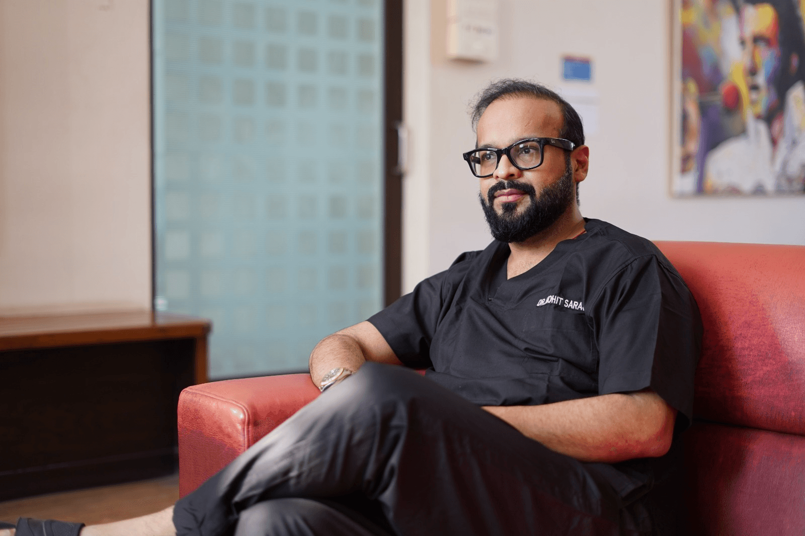 dr moht - Saraogi Hospital Man with beard and glasses in black scrubs sitting on a red sofa in a clinic lobby/ waiting area. Care professional relaxing in reception area.